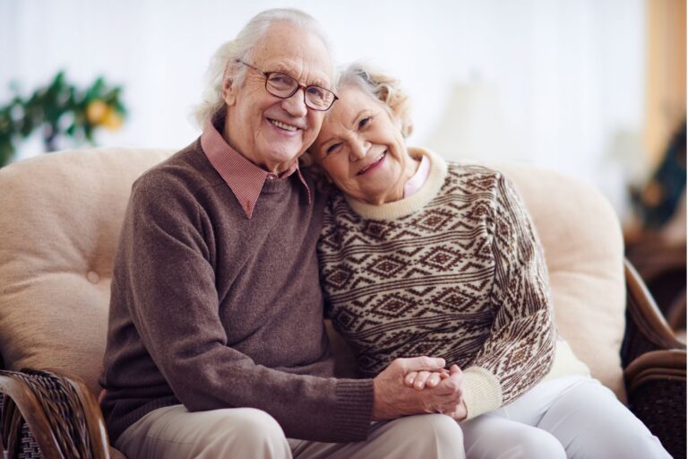 An image of an elderly couple, sat on a sofa holding hands.