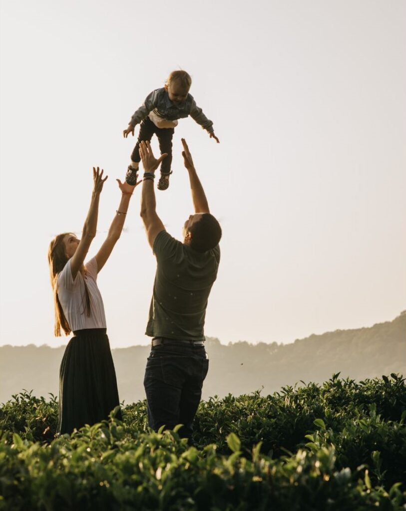 A couple are playing with their baby in nature. The baby is thrown up and caught by the dad.